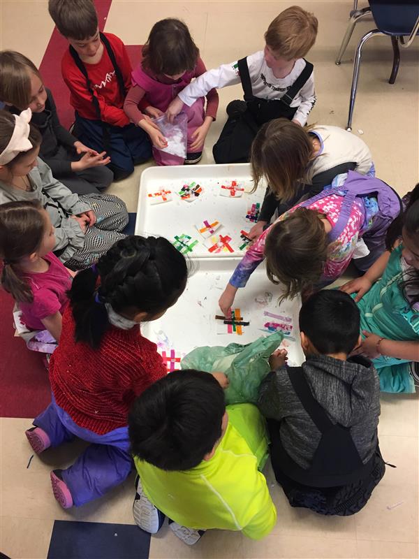 This image shows students experimenting with snow to make art using tissue paper that bleeds when it gets wet. Students are all gathered around a big tray watching the snow melt. As the snow melts, the artwork emerges.